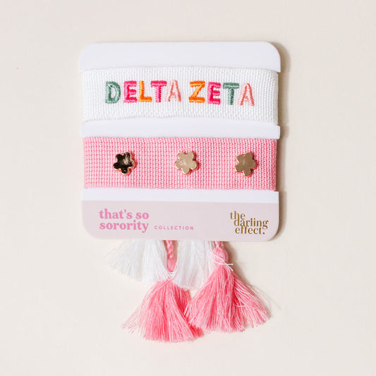 The Darling Effect Delta Zeta Woven Bracelet Set features two adjustable bracelets—one white with DELTA ZETA in multicolored letters, one pink with gold flower studs—both accented with pink and white tassels. Ideal as a sisterhood gift.