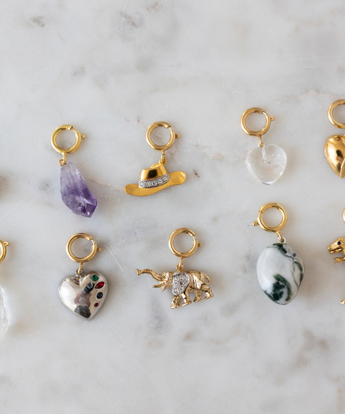 Nine gold jewelry charms—featuring shapes like hearts, stones, a cowboy hat, and an elephant—are displayed on marble as the perfect inspiration for your next DIY Schmuck-Workshop x MINI im KaDeWe Berlin by WALD Berlin or your Charm DIY project.