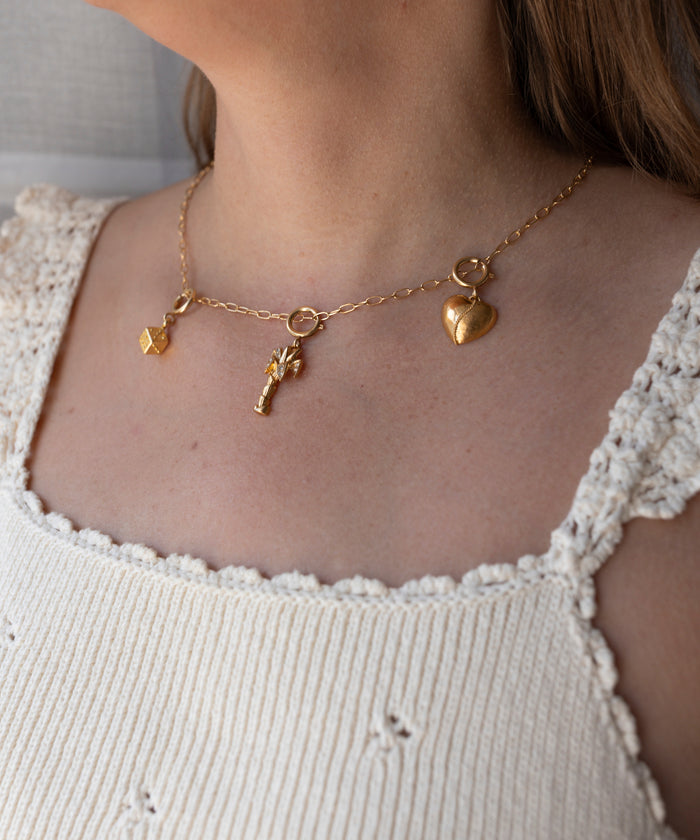 A gold chain necklace by WALD Berlin featuring three charms—a cube, a key, and a heart—styled atop a white crocheted tank top. This piece is inspired by the DIY Schmuck-Workshop x MINI at KaDeWe Berlin, bringing handcrafted elegance to any look.