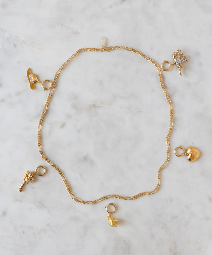 Showcased on a light marble surface, this gold chain necklace by WALD Berlin features five assorted gold charms—a shoe, lock, heart, animal, and floral shape—capturing the playful essence of the brand and offering a unique opportunity for personalization at the DIY Schmuck-Workshop x MINI im KaDeWe Berlin.