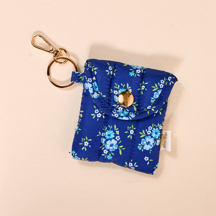 The-darling-effect's Endless Daydream Navy Puffy Earbud Case Keychain features a quilted floral design, gold snap button, and keyring clasp—a stylish wireless earbud case and charming accessory, shown on a beige background.