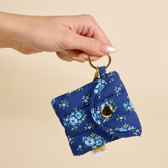 A hand with manicured nails holds the-darling-effect Endless Daydream Navy Puffy Earbud Case Keychain—a small, quilted pouch with a blue floral pattern and button closure—by a gold keyring against a beige background.