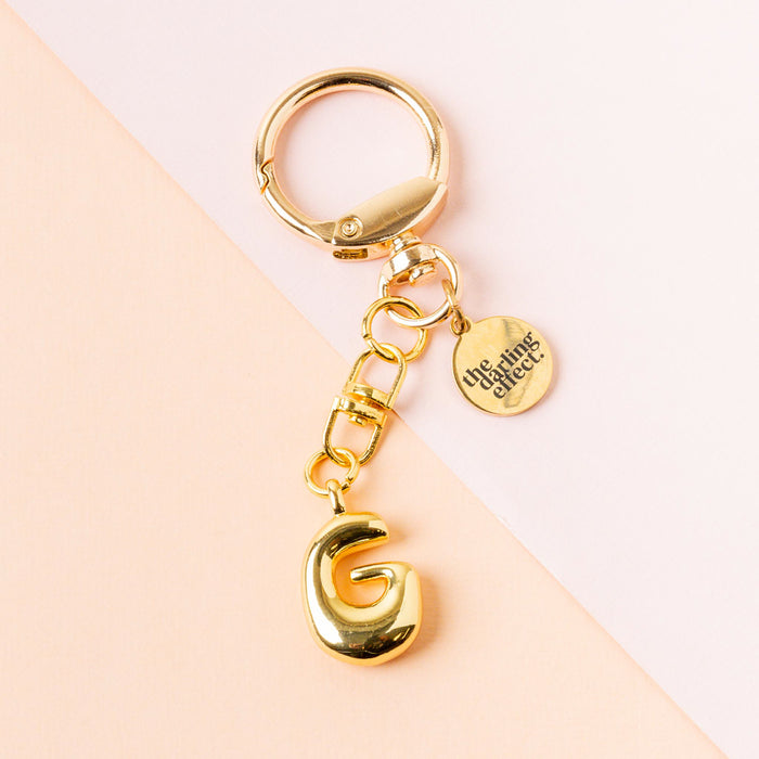 The G Bubble Letter Keychain Bag Charm by the-darling-effect features a bold gold bubble letter G and a round tag engraved with The Aimes Effect, displayed on a soft peach and pale pink background.