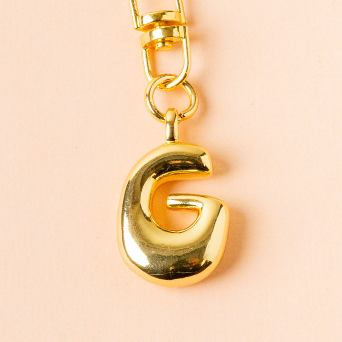 The G Bubble Letter Keychain Bag Charm by the-darling-effect features a shiny gold G pendant on a gold chain set against a light peach background, making it an ideal personalized keychain or stylish bag charm.