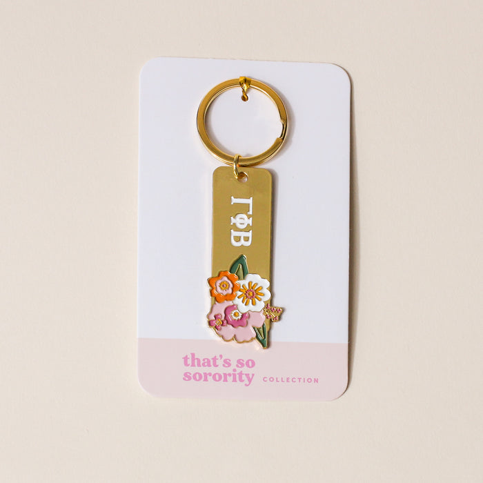 The Darling Effect's Gamma Phi Beta Enamel Keychain, featuring Greek letters and colorful enamel flowers, is displayed on a white card labeled "that's so sorority collection" in pink—perfect for any sorority gift.