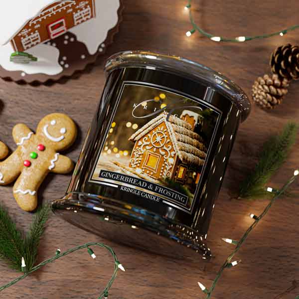 Decorative candle with gingerbread house design on a wooden surface with cookies and lights.