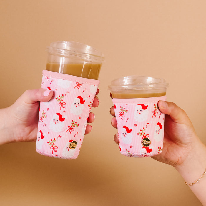 Two hands hold iced coffee cups with The Darling Effect's Holly Jolly Santa Sip Savvy Drink Sleeves, featuring Santa faces, holly, and bows against a beige background.