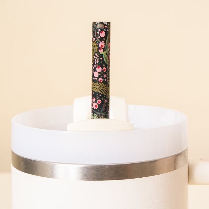 A close-up of The Darling Effect’s Jolly Sprig Green 40 oz Tumbler Straw Set, showcasing a white tumbler with a metallic rim and an eco-friendly black straw adorned with a colorful floral and cherry design, set against a light beige background.
