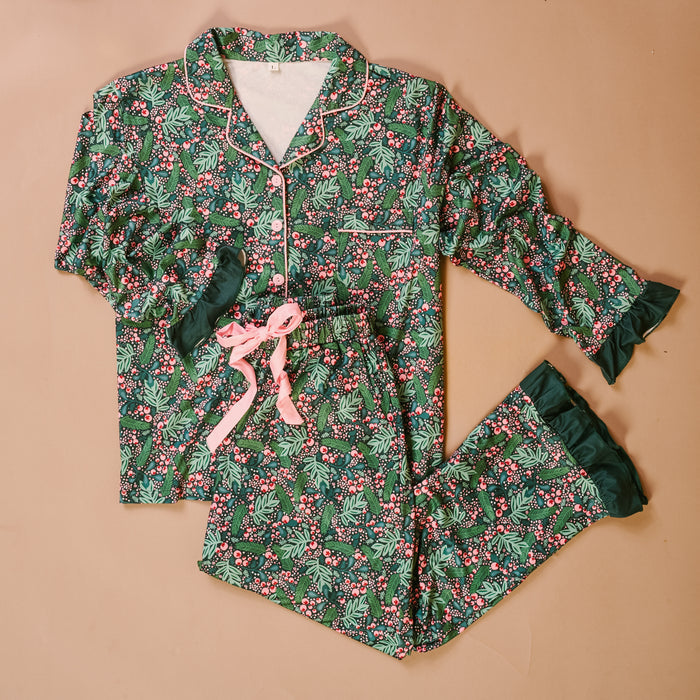 The Jolly Sprig Sweet Dreams Pajama Pants Set by The Darling Effect features a green background adorned with a festive holly and berry pattern, accented with pink piping, a matching bow, and ruffled cuffs on both the sleeves and pants. Crafted from ultra-soft fabric for ultimate comfort, this cozy pajama set is displayed laid flat against a neutral background.