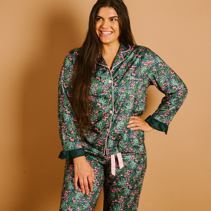 A woman stands against a tan background, smiling as she poses with one hand on her hip, wearing The Darling Effect’s Jolly Sprig Sweet Dreams Pajama Pants Set—matching green floral pajamas with pink accents and white piping crafted from ultra-soft fabric—perfectly embodying the comfort and charm of cozy pajama sets.