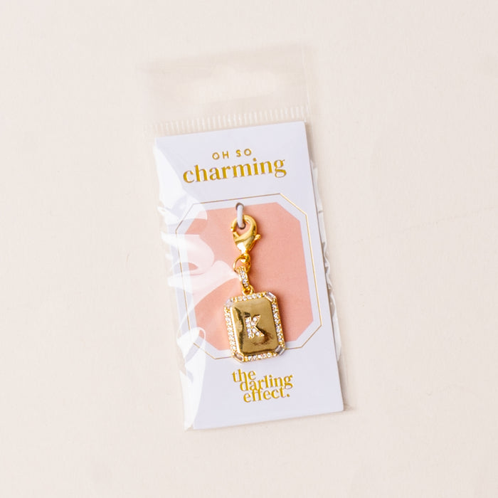 The K Jewelry Charm Initial Letter Gold by the-darling-effect features gold plating and rhinestones, attached to a lobster clasp. It comes in a clear package labeled "oh so charming" with a light neutral background for a personalized touch.