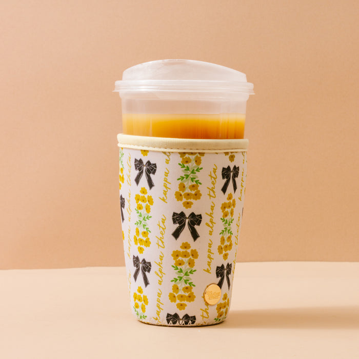 A clear plastic cup with orange juice sits inside The Darling Effect's Kappa Alpha Theta Large Sip Savvy Drink Sleeve, featuring yellow flowers, black bows, and cursive on beige—perfect for stylish sorority gifts.