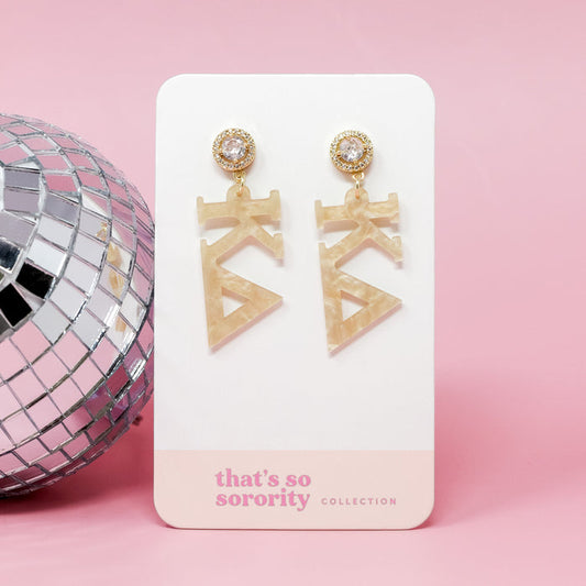 The Darling Effect’s Kappa Delta Letter Gold Acrylic Earrings feature clear gems and cream Kappa Delta letters, displayed on a white card next to a disco ball—lightweight and perfect for any bid-day basket.