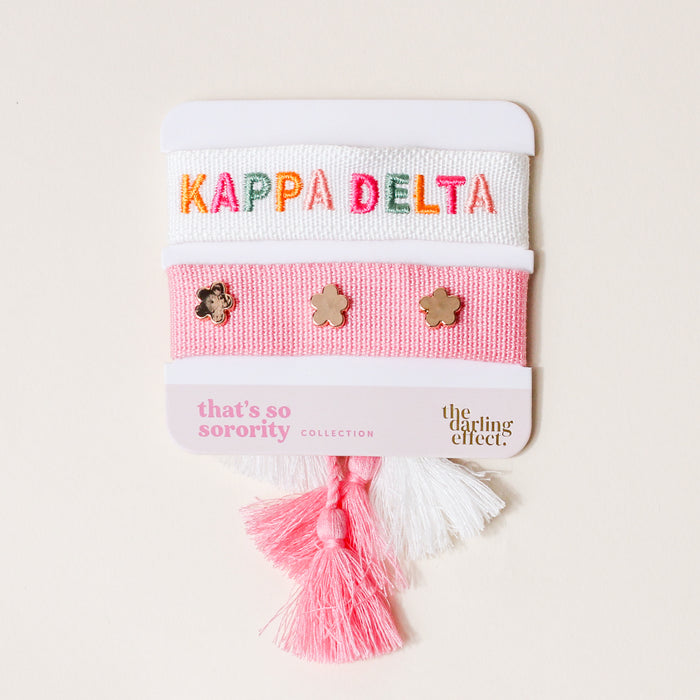 The Darling Effect's Kappa Delta Woven Bracelet Set features two bracelets—one white with colorful KAPPA DELTA text, one pink with gold flower charms—plus pink and white tassels. A great sisterhood gift from the "that's so sorority" collection.