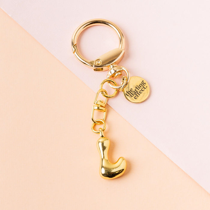 The-darling-effect's L Bubble Letter Keychain Bag Charm features a gold finish and a round tag engraved with "the gifting affair," perfect as a personalized bag charm, shown on a soft pink and peach background.
