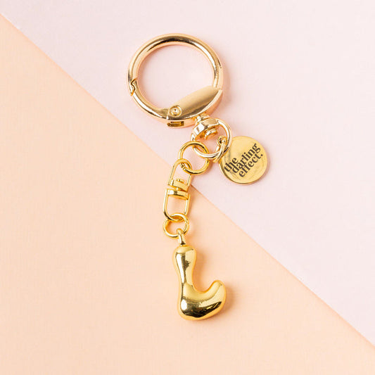 The-darling-effect's L Bubble Letter Keychain Bag Charm features a gold finish and a round tag engraved with "the gifting affair," perfect as a personalized bag charm, shown on a soft pink and peach background.