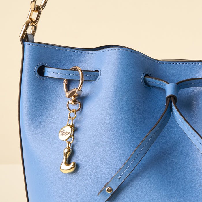Close-up of a light blue leather handbag with a drawstring closure, gold chain, and two gold charms—a small disc and a shoe—on the front. The-darling-effect’s L Bubble Letter Keychain Bag Charm adds flair against the plain, neutral background.