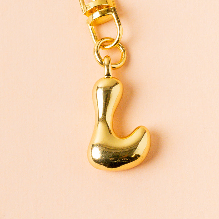 The-darling-effect's shiny, gold L Bubble Letter Keychain Bag Charm hangs from a clasp against a light peach background.