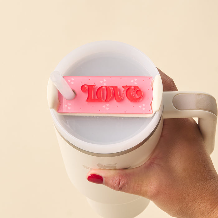 A hand with red nail polish holds a white mug featuring The Darling Effect’s Love Tumbler Lid Tag—a pink chocolate-shaped accessory with "LOVE" and hearts—that adds personalized charm to your drinkware.
