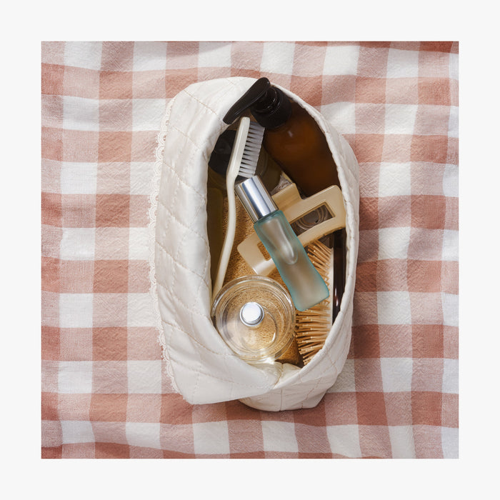 Cream | Top view of open Monos x Reformation cosmetic pouch in Cream filled with beauty products on a checkered picnic blanket