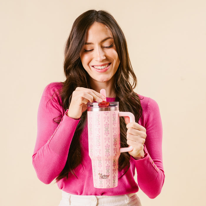 A smiling woman in a pink top holds the-darling-effect's Oh So Fancy Pink 40 oz Lifestyle Flip Straw Tumbler with both hands, stirring her drink with the straw. The background is plain and light-colored.