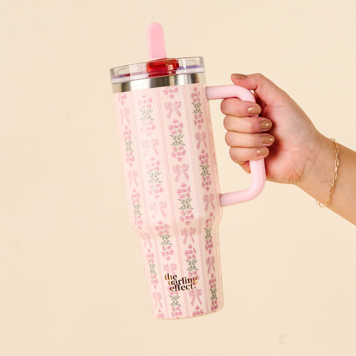 A hand holds the-darling-effect’s Oh So Fancy Pink 40 oz Lifestyle Flip Straw Tumbler, featuring floral and bow designs, a handle, clear lid, pink flip straw, and leak-resistant style—perfect for practical on-the-go sipping.