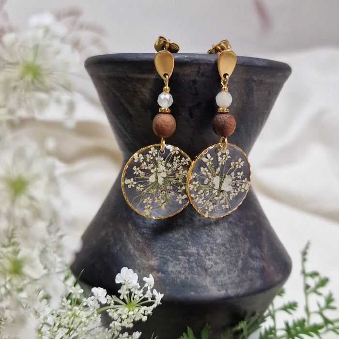Handgefertigte Ohrringe 'Perlblumen' mit Nussholz, kristallklarem Resin mit weißen Gierschblüten und Süßwasserzuchtperlen auf dunklem Dekoelement.