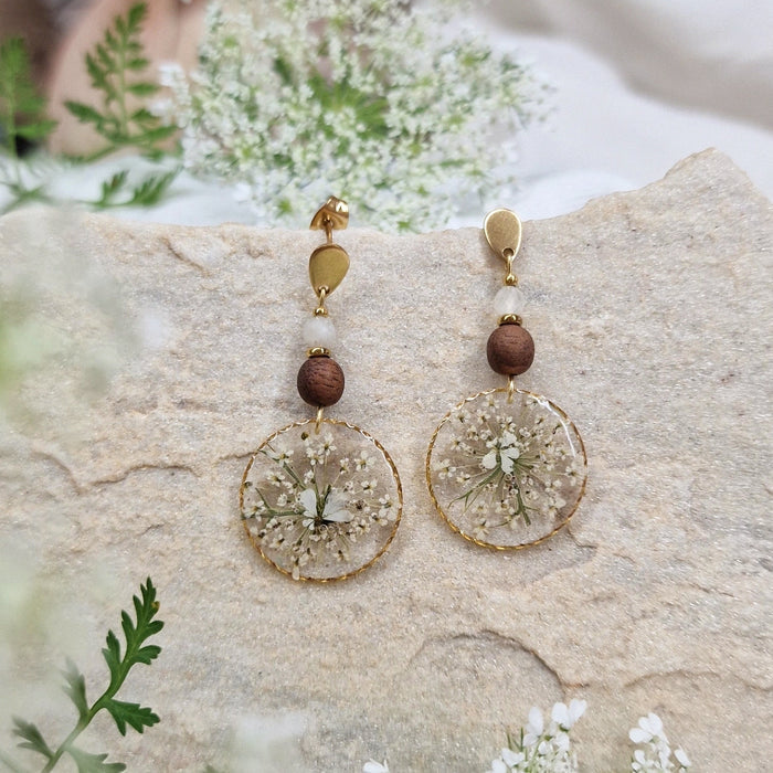 Handgefertigte Ohrringe mit Nussholzscheibe, transparentem Resin mit getrockneten weißen Gierschblüten und Perle auf hellem Naturstein.