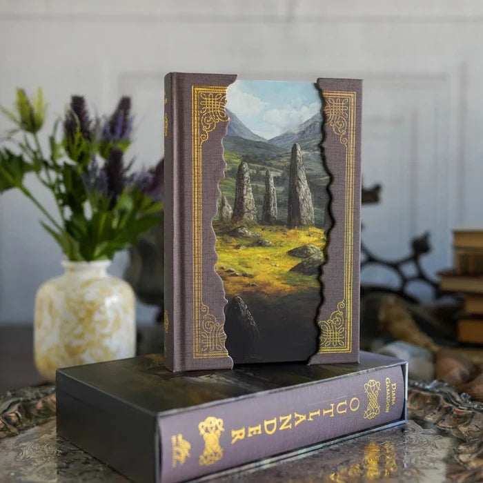 Outlander Annotated Special Edition hardcover book displayed upright on a dark decorative surface, featuring a stunning cover art of Scottish Highland standing stones with dramatic mountains and green fields framed by ornate gold borders, with a lavender floral arrangement in the background