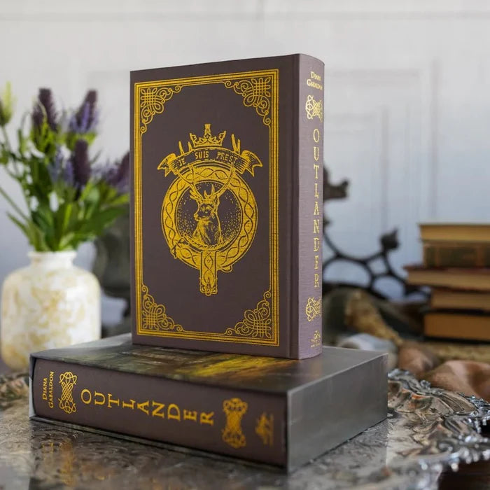 Outlander Annotated Special Edition hardcover book showing the Fraser clan crest cover with gold "Je Suis Prest" motto, stag emblem, and ornate gold border on a dark brown background, displayed upright on its slipcase with a lavender floral arrangement in the background