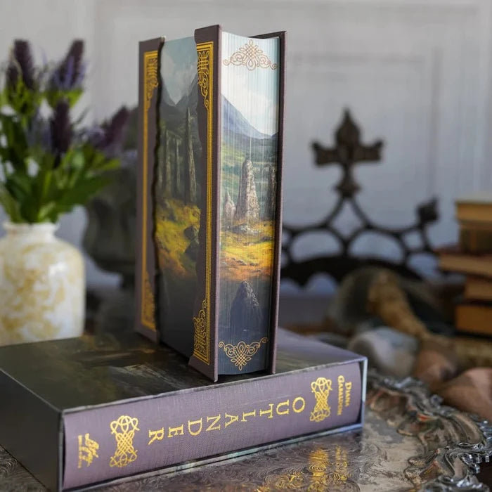 Outlander Annotated Special Edition hardcover book being slid out of its decorative dark brown slipcase, revealing the Scottish Highland standing stones cover art with gold borders, displayed on the slipcase base which shows "Outlander Diana Gabaldon" in gold lettering