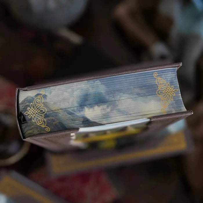 Close-up of the Outlander Annotated Special Edition fore-edge showing a hand-painted Scottish Highland mountain landscape with dramatic cloudy skies on the gilded page edges, accented with gold Celtic knot ornaments at the corners, a premium special edition feature