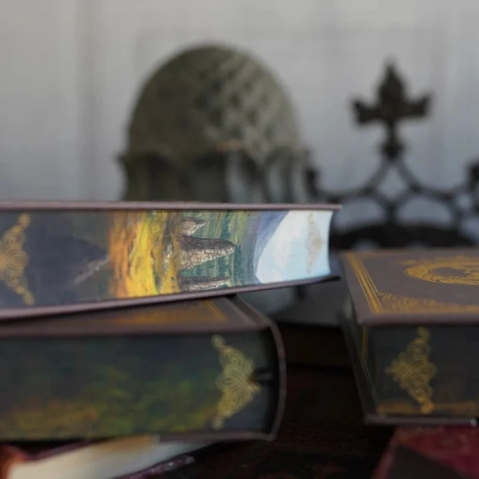 Side view of the Outlander Annotated Special Edition hardcover book and slipcase stacked together, both displaying matching painted fore-edge artwork of Scottish Highland standing stones with golden fields and dramatic sky, with decorative sculptural pieces in the blurred background