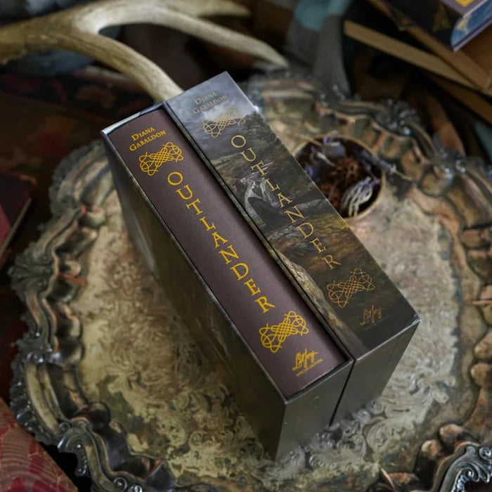 Overhead flatlay view of the Outlander Annotated Special Edition hardcover book being removed from its dark brown slipcase, showing both spines with gold "Diana Gabaldon Outlander" lettering and Celtic knot designs, with an author signature at the base, displayed on a decorative tray surrounded by antlers