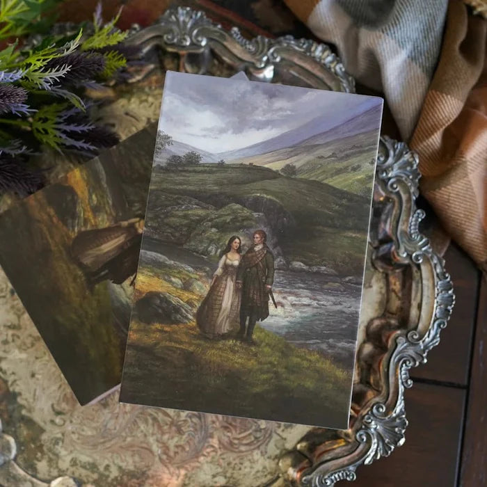Two Outlander Annotated Special Edition illustrated slipcases displayed on an ornate silver decorative tray, showing the exterior slipcase artwork featuring a romantic Scottish Highland river scene with Jamie and Claire Fraser (front) and a second slipcase behind it, with tartan fabric and Scottish thistles in the background