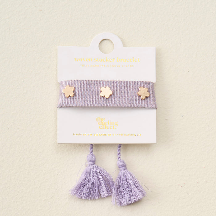 The Darling Effect’s Periwinkle Woven Stacker Bracelet features two tassels and three gold flower charms, displayed on a white card with gold text.