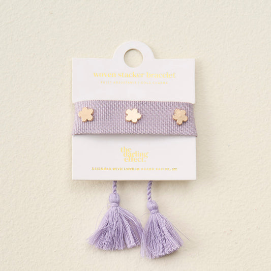 The Darling Effect’s Periwinkle Woven Stacker Bracelet features two tassels and three gold flower charms, displayed on a white card with gold text.