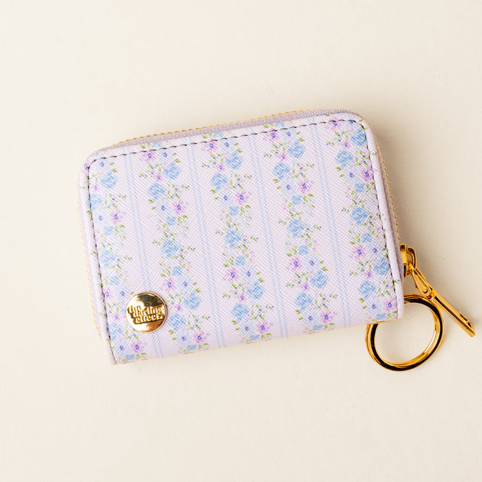 Introducing the Petal Parade Blue Zip Around Wallet from the-darling-effect—a compact and stylish accessory in soft blue. This keyring wallet showcases a delightful floral pattern and gleaming gold zipper closure. Multiple card slots keep your essentials organized, while a round gold emblem on the bottom left corner adds an elegant finishing touch.