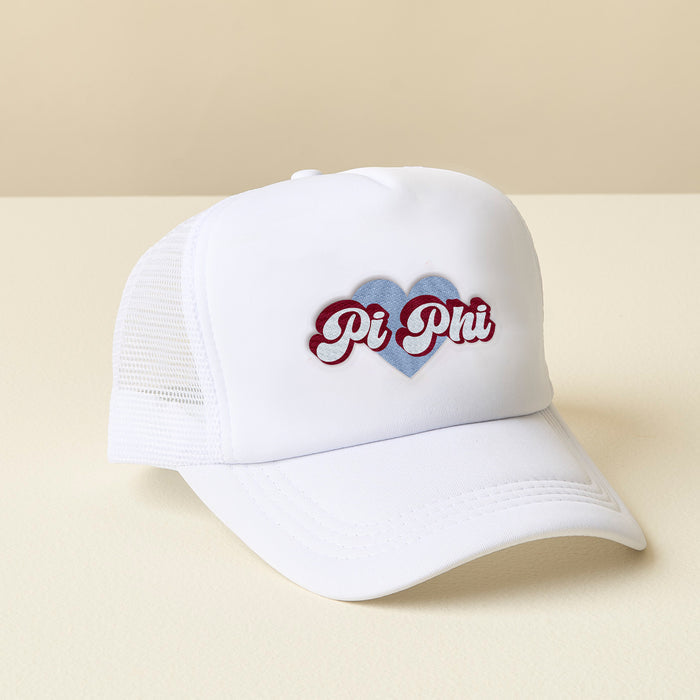 The Darling Effect’s Pi Beta Phi White Trucker Hat features red script over a blue heart on the front panel—shown on a cream surface with a beige backdrop—making it an ideal sisterhood gift.
