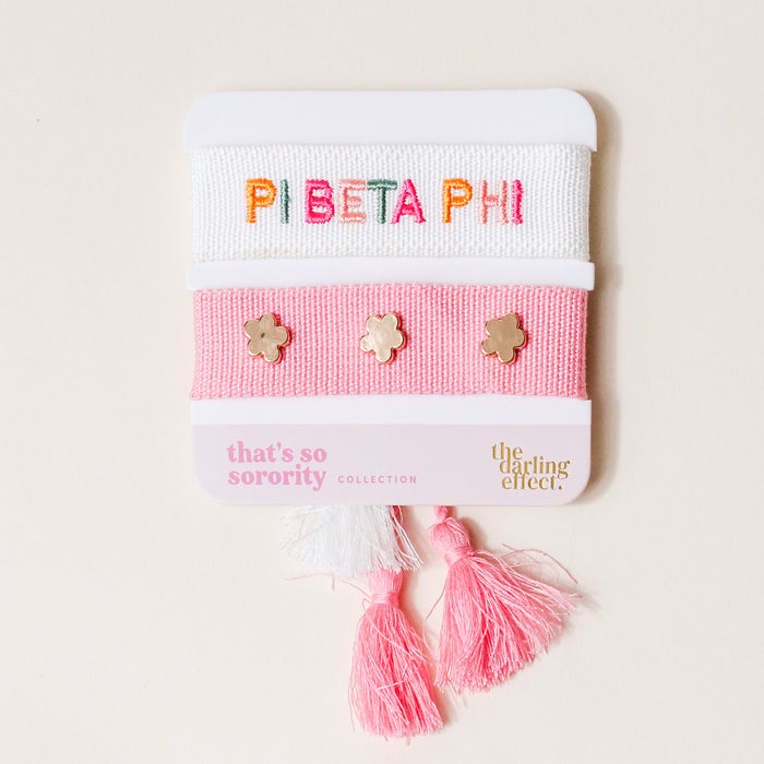 The Darling Effect Pi Beta Phi Woven Bracelet Set features two bracelets—a white one with colorful “PI BETA PHI” embroidery, and a pink one with gold flower studs—plus pink and white tassels, creating an ideal sorority gift or sisterhood accessory.