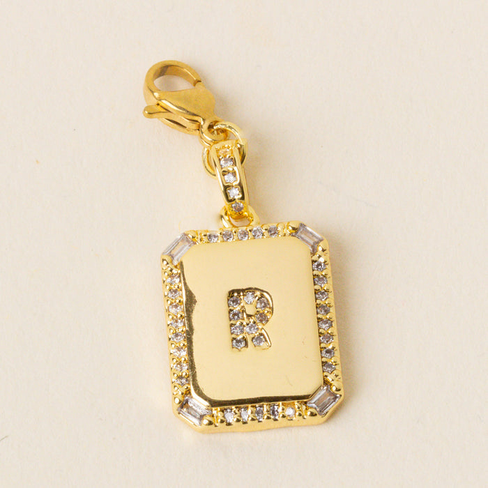 The-darling-effect’s R Jewelry Charm Initial Letter Gold features a gold-plated rectangular pendant with a diamond-studded border and diamond-encrusted cutout R, attached to a gold lobster clasp for easy personalization.