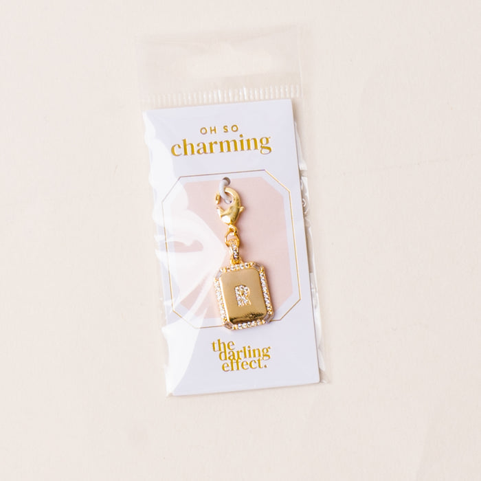 The R Jewelry Charm Initial Letter Gold by the-darling-effect features a gold-plated rectangular pendant with an 'R' in crystals, surrounded by sparkling stones and a lobster clasp. Packaged with signature Oh So Charming branding.