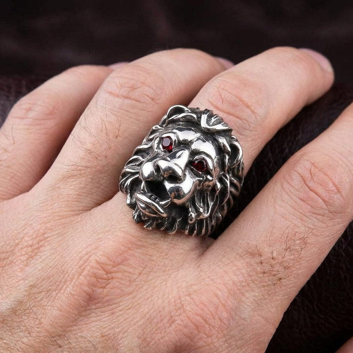 Sterling silver lion head ring with red gemstone eyes worn on a man's finger, featuring detailed mane carving and oxidized finish in a bold biker style design