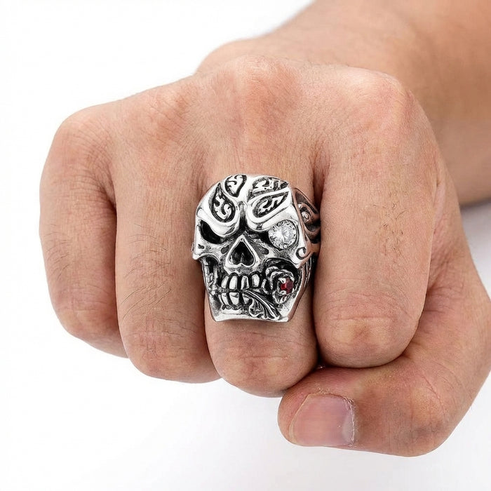 Close-up of a hand wearing a detailed silver deadly rose skull ring. The gothic biker ring features tribal engravings, a clear gemstone eye, and a rose with a red stone in its mouth, modeled on a clenched fist against a white background.