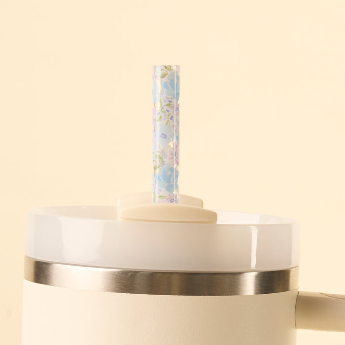 A close-up of the-darling-effect’s Secret Garden Blue 40 oz Tumbler Straw Set, a white tumbler with metal rim and BPA-free reusable clear straw featuring a pastel floral pattern, shown against a pale beige background.