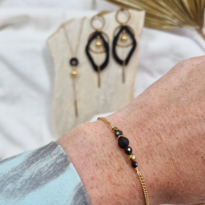 Handgefertigtes Armband aus dunklen Holzperlen mit goldfarbener Kette am Handgelenk getragen, im Hintergrund passende Ohrringe und Halskette
