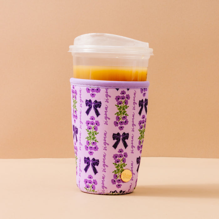 The Darling Effect Sigma Sigma Sigma Large Sip Savvy Drink Sleeve wraps a clear plastic cup with an orange drink, featuring purple bows, green floral sprigs, and repeating "hygge" in purple on a light beige background.