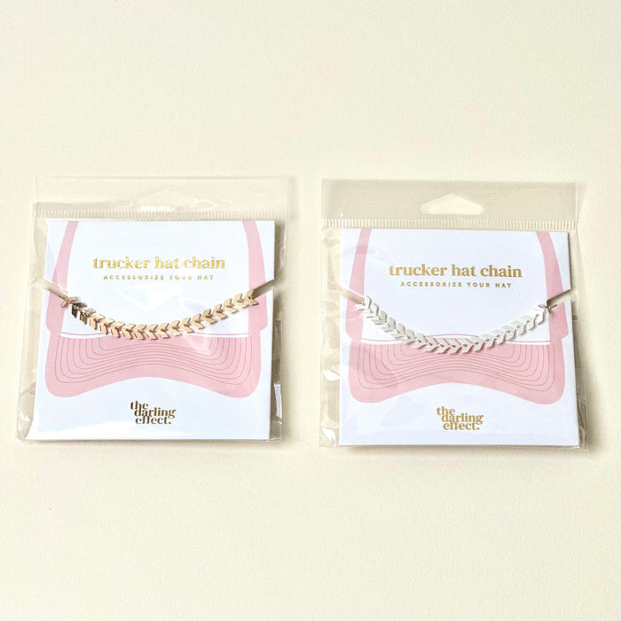 Two Silver-Leaf Chain Trucker Hat Chains by The Darling Effect are packaged on cards with pink wavy lines and the brand logo. One accessory is gold, the other silver—both add flair to mesh-back caps with chic chevron designs.