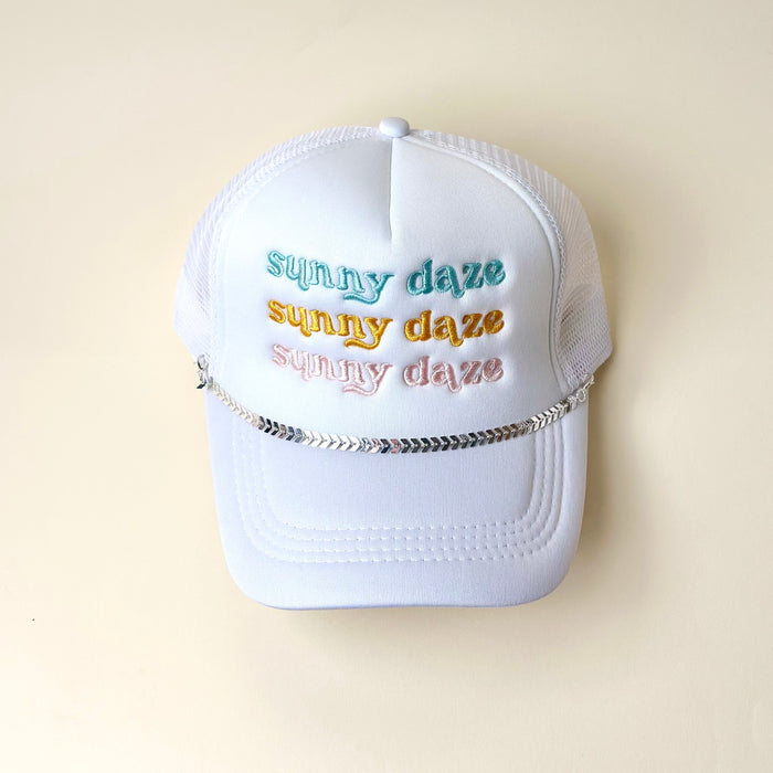 A white trucker hat with mesh back features "sunny daze" embroidered in blue, yellow, and peach. The Darling Effect's Silver-Leaf Chain Trucker Hat Chain decorates the front above the brim. Background is off-white.