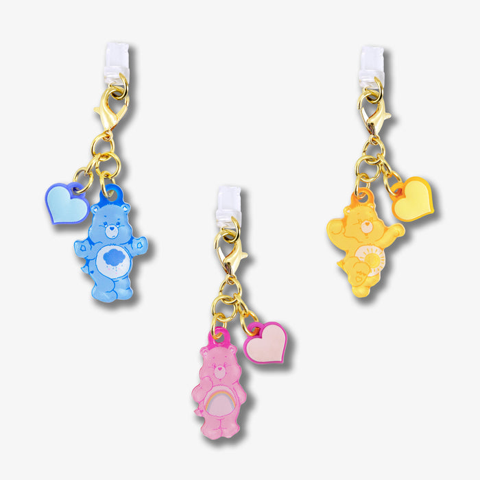 Close-up of Care Bears pastel port charms 3-pack showing gold lobster clasps and white silicone plugs on white